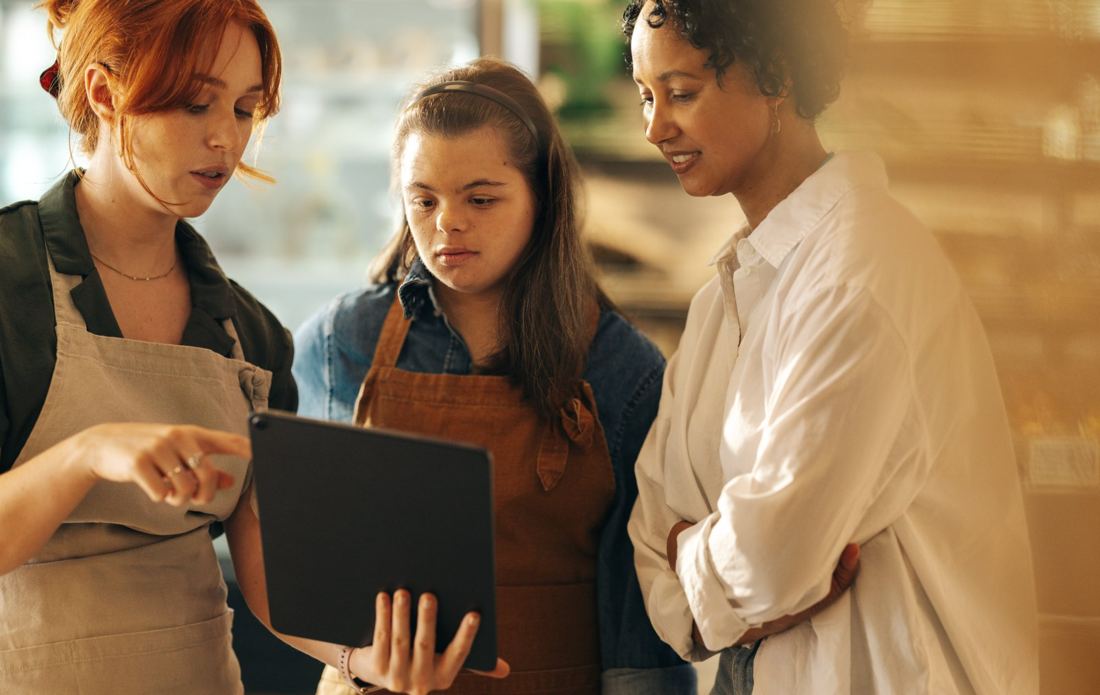 A worker in an apron points at a tablet held by a young woman with Down syndrome in a café, while a third colleague looks on.